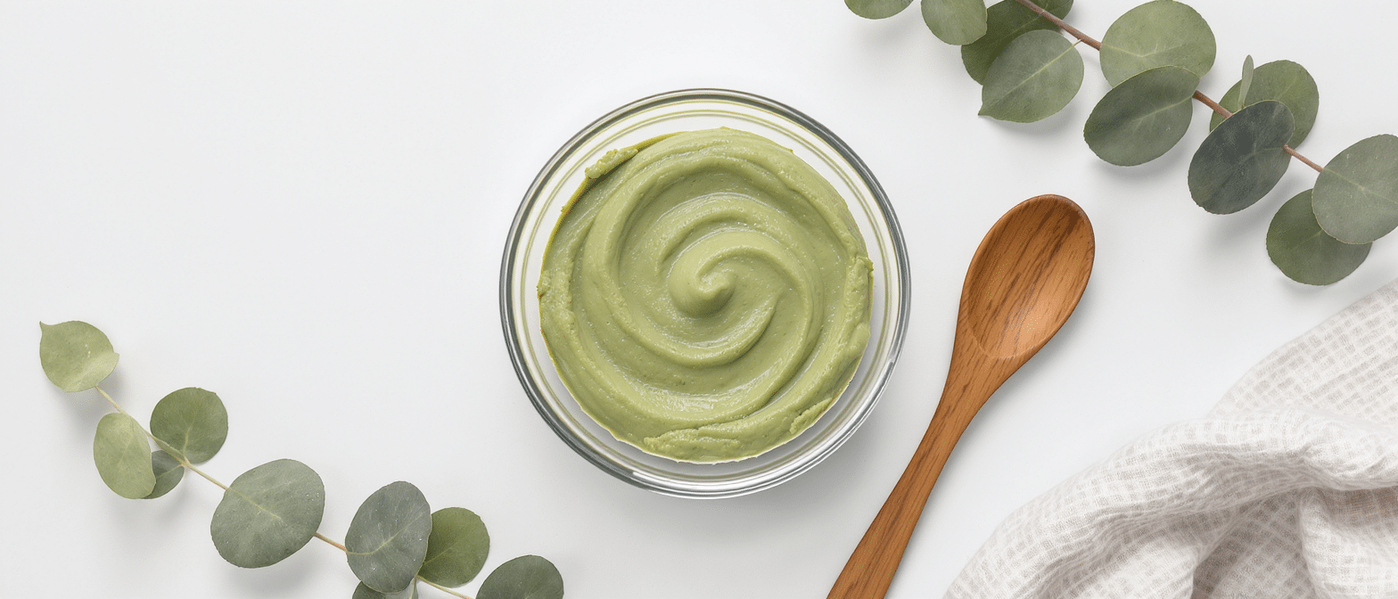 Bol de masque à l’argile verte prêt à l’emploi, entouré de feuilles d’eucalyptus et d’une cuillère en bois sur fond blanc.